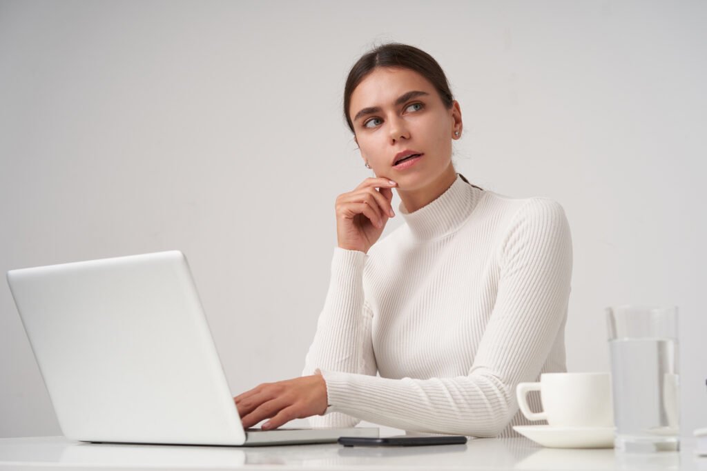 Puzzled young beautiful dark haired lady with natural makeup touching her face with raised hand and looking thoughtfully, working at modern office with laptop over white background